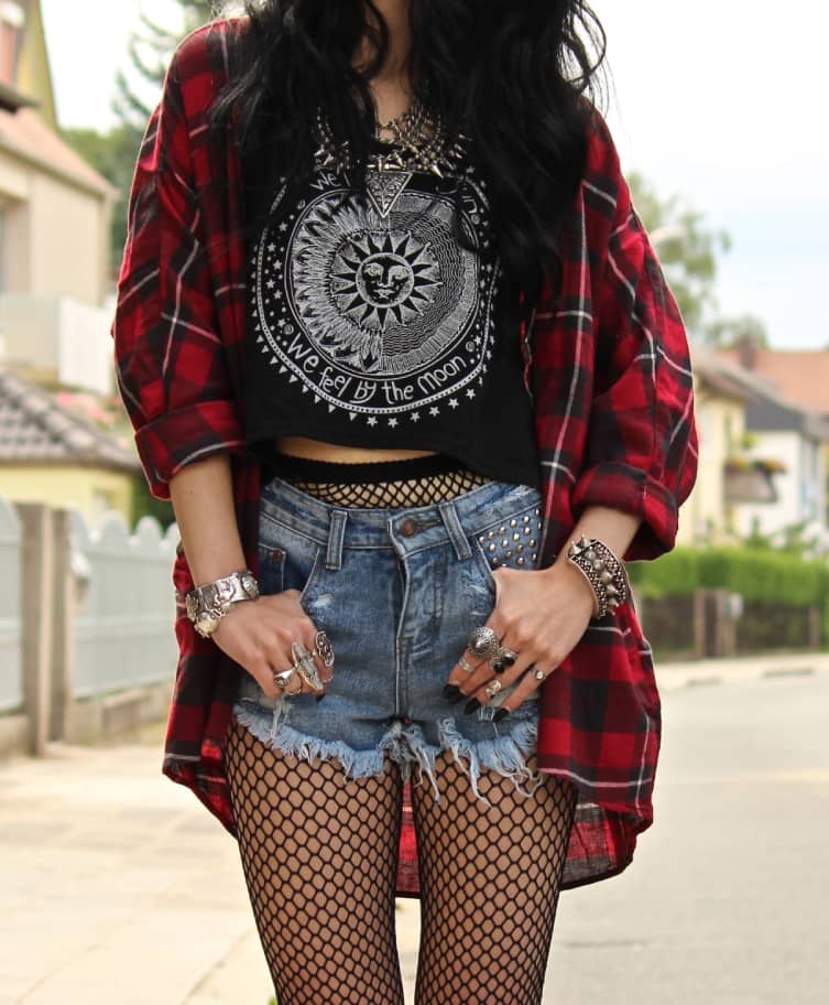 Woman wearing a black graphic tee, distressed denim shorts, fishnet stockings, and an oversized plaid shirt, accessorized with silver rings and bracelets.